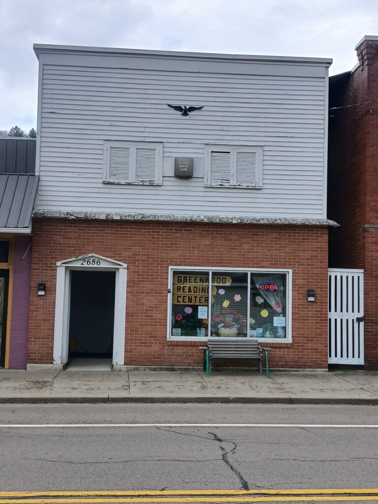 Image of the Reading Center Building; a brick building with a white door frame and window, and a white, wooden second floor. An 'open' sign shines in the upper right corner of the window, while a wooden sign on the left reads 'Greenwood Reading Center.' The window is decorated brightly with books and flowers.