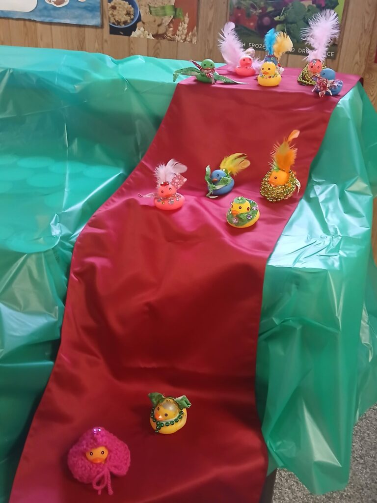Image of a red, silk runner draped over a table. A series of rubber ducks are clustered in various places on the runner. Each duck is decorated with beads, cloth, sequins and feathers.